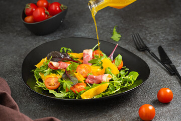 Fresh salad with arugula, cherry tomatoes, orange slices and smoked duck slices on a black plate on a dark concrete background. Served with olive oil dressing. Salad recipes.