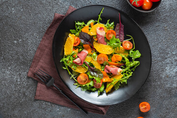 Fresh salad with arugula, cherry tomatoes, orange slices and smoked duck slices on a black plate on a dark concrete background. Served with olive oil dressing. Salad recipes.
