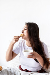 Young brunette woman sitting in the bed with eating croissants