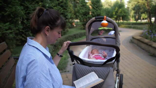 A young mother sways the baby in a stroller with one hand. Girl is reading a book sitting on a park bench. 4K Slow Mo