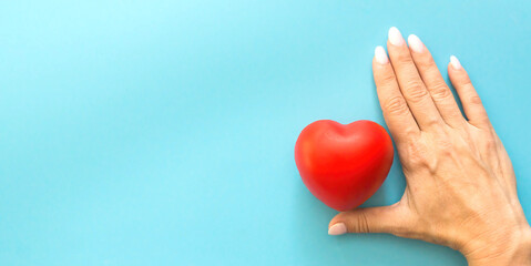 a female hand protects a red toy heart on a blue background, a banner on the left copyspace,...