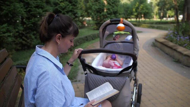 A young mother is reading a book, a baby is being played in a stroller. 4K Slow Mo