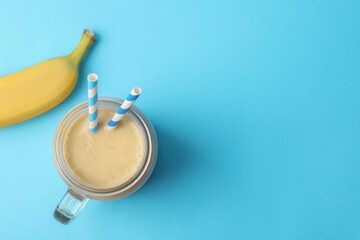 Mason jar of tasty banana smoothie with straws and fresh fruit on light blue background, flat lay. Space for text