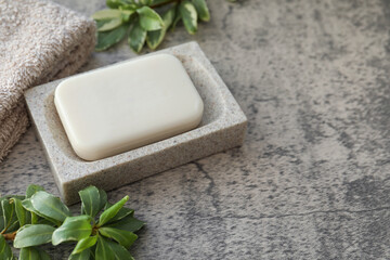 Dish with soap bar, terry towel and green plants on light grey textured table. Space for text