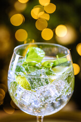 Transparent cocktail drink in glass with ice, and lime on a dark rustic board in Christmas decoration with Christmas tree and lights in blurry in background. Gin and tonic or cocktail called Hugo