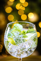 Transparent cocktail drink in glass with ice, and lime on a dark rustic board in Christmas decoration with Christmas tree and lights in blurry in background. Gin and tonic or cocktail called Hugo