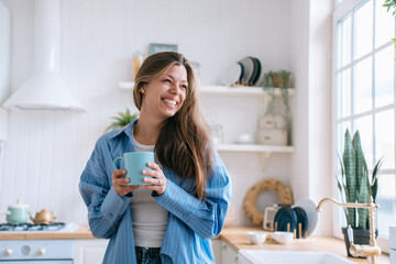 Happy caucasian female with long loose hair in jeans and blue shirt toothy smiles holds cup of tea...