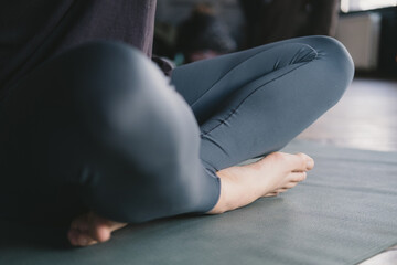Close-up photo of the legs of a female yogi meditating barefoot and wearing leggings in easy pose on a yoga mat on the floor