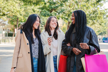 Three multiracial happy young women walking and talking on a shopping day. Group of girls carrying bags smiling on the street on a sale week. Consumism concept. High quality photo