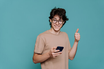 Handsome teenager in glasses and beige t-shirt holds phone shows thumb up gesture looks at camera...