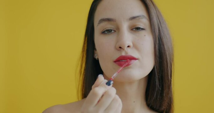 Close-up slow motion portrait of young lady applying lipstick then sending air kiss on yellow background. Beauty and decorative cosmetics concept.