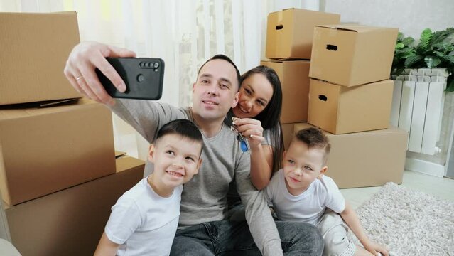 Excited parents with children enjoy moving into new apartment. Family with happy expressions makes selfie with keys against unpacked carton boxes