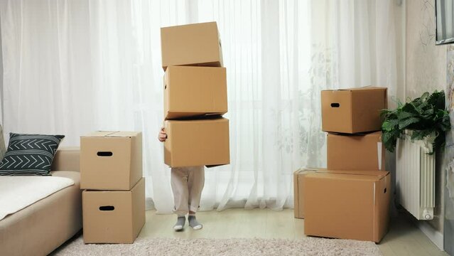 Preschooler boy tries to carry boxes stack in new apartment and struggles with holding in arms. Carton boxes hide kid falling on floor and child looks around