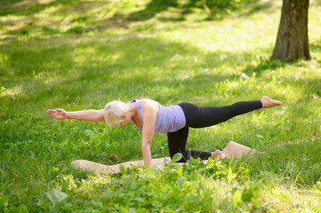 Full length portrait of a middle aged woman doing yoga or pilates on a mat outside in a park.Pose chakravakasana.