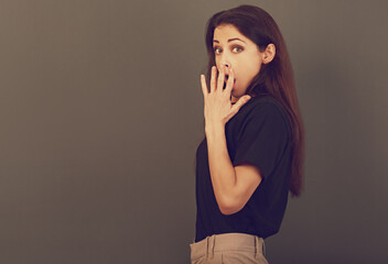 Surprised beautiful woman looking with big eyes, opened mouth and covering mouth  the hand in black shirt on grey background. Closeup