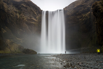 skógafoss, la cascata in lunga esposizione a formare una tenda d'acqua con visibile il fiume e i sassi su cui c'è la sagoma di un uomo solo