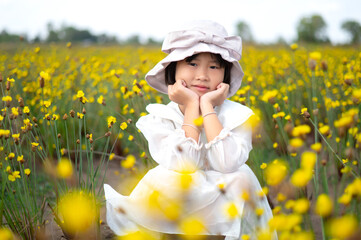 Cute Japanese,Korean Girl Kid  was wearing a white dress and a hat.Sitting in a field of bright yellow flowers, very beautiful.There is blur and bokeh in the foreground and background.