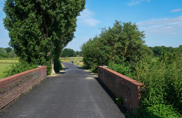 Neu angelegter Radweg mit frisch asphaltierter Fahrbahndecke in einer idyllischen Natur im Sommer mit einem Radfahrer im Hintergrund und einer Brücke im Vordergrund