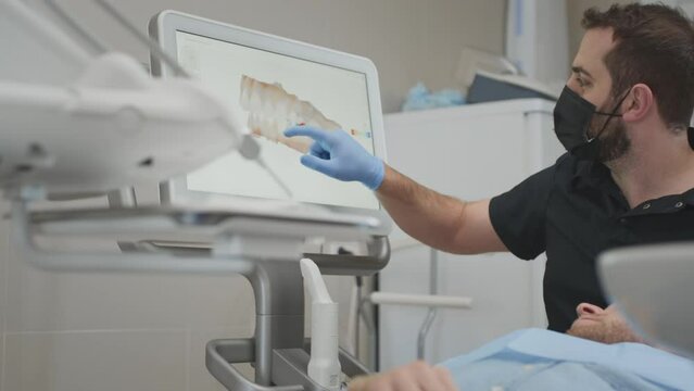 A male orthodontist doctor shows on the display a 3d model of the oral cavity of a man patient. Medical examination of teeth. Digital scanning with a scanner, checking the jaw. Dental clinic office.