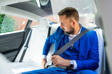 A caucasian man in a blue suit fastens his seat belt in the back seat of a car. Business class passenger. 