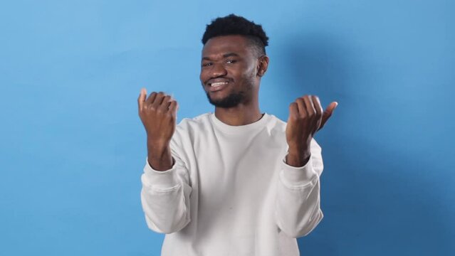 A handsome African man calls Hey you come here. The guy asks to join him, beckons with an inviting gesture the blue studio background.