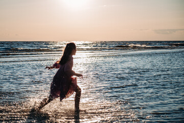 Silhouette of teen girl with loose hair and wearing airy dress running along the beach with water splashes at golden hour. Focus on water