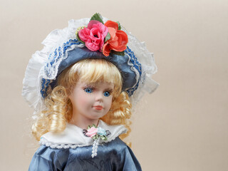 Vintage porcelain doll baby girl with blue eyes, blonde curls, wearing a blue hat decorated with red flowers and a blue satin dress.