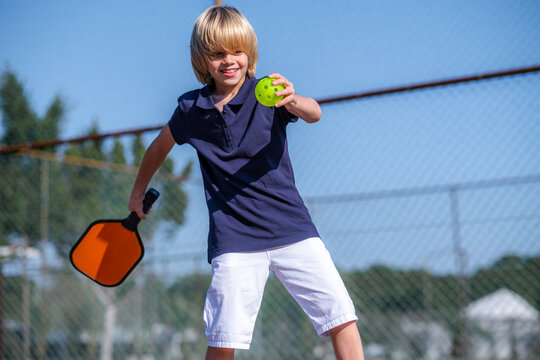 Happy blonde boy playing pickleball game, hitting pickleball yellow ball with paddle, outdoor sport leisure kids activity.