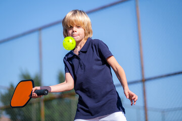 Happy blonde boy playing pickleball game, hitting pickleball yellow ball with paddle, outdoor sport...