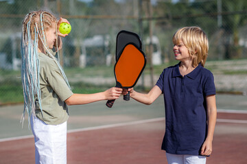 Laughing boy and girl playing pickleball game, hitting pickleball yellow ball with paddle, outdoor...