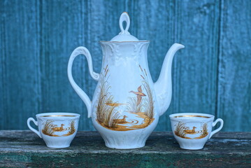 a service of a white ceramic teapot and two cups with a brown pattern stands on a gray table against a green wall