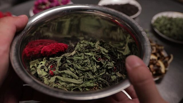 The compiler of the tea composition adds berries, dried flowers and other ingredients to the dry tea leaves lying in a metal bowl.