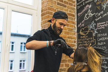 Fototapeta na wymiar Absorbed hairdresser in the black hat with beard styling his client's long hair in the professional hair salon. . High quality photo