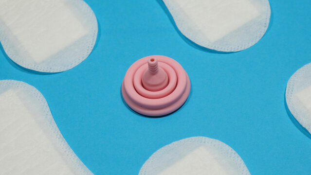 Copa menstrual rosa de l&aacute;tex plegada entre compresas en un fondo azul. Menstruaci&oacute;n sostenible