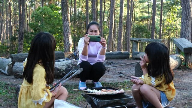 Happy mother taking pictures of her adorable daughter having a picnic by the stove near the tent and grilling in the pine forest. Happy family on vacation in nature.