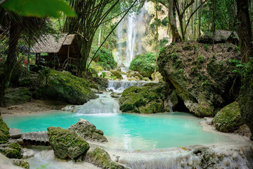 A clearing in the tropical jungle with a turquoise pond in the foreground and many moss-covered...