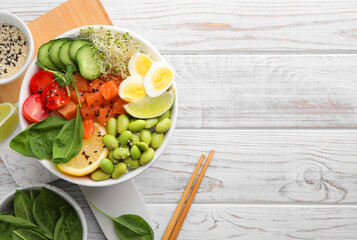 Delicious poke bowl with quail eggs, fish and edamame beans on white wooden table, flat lay. Space...
