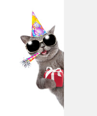 Happy cat wearing sunglasses holds gift box, blows in party horn and looks from behind empty white banner. isolated on white background
