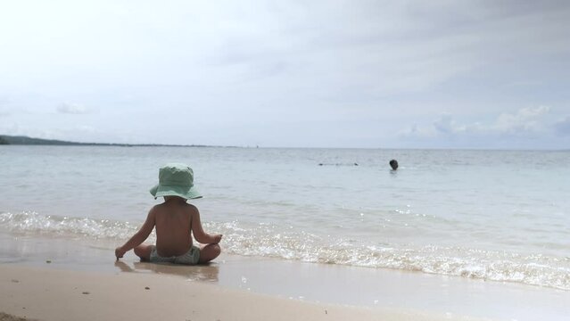 A small child in a children's panama sits on the seashore. Sea waves wash the baby's legs. Swimming in warm water on a hot summer day on Bohol island. The best family vacation in an exotic location.
