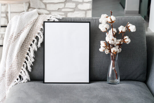 Mock up of black frame with white background. Cotton flowers and empty vertical template on sofa. Cozy living room interior. Home decorations on couch and plaid. Copy space