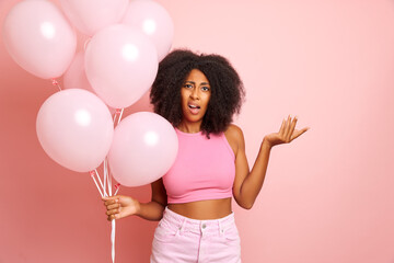 Mixed race woman with complaint face, holds bunch of pink balloons with unexpected surprise emoution, isolated over pink wall