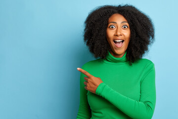 Indoor shot of positive curly haired woman with overjoyed expression, points index finger and shows copy space for your advertisement, wears green jumper, poses over blue wall