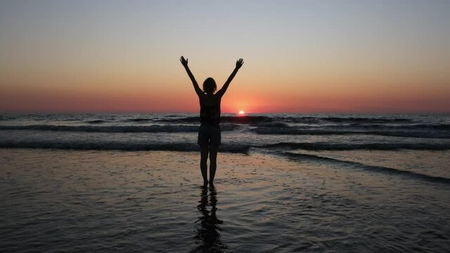 Happy woman raises her hands up on the ocean background. Silhouette of a young female tourist walking relaxed on the sea at sunset.