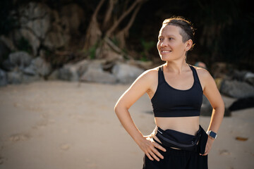 Fototapeta premium Female athlete on a tropical beach in a sports top and black shorts rests from exercise and looks away.
