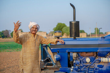 indian senior farmer standing with tractor at agriculture field.