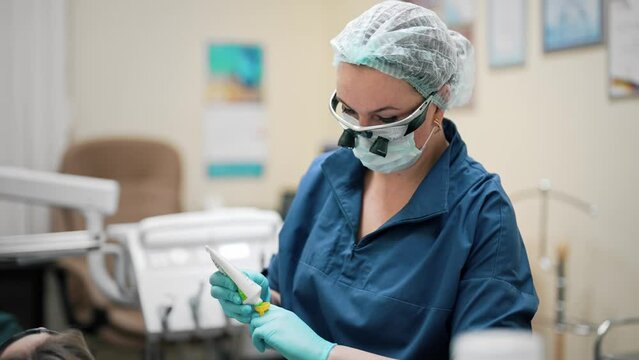 Woman lying on chair on cabinet tooth clinic with mouth dilator and protective goggles. Dentist in glasses with magnifying glass holding medical equipment. UV lamp for whitening or caries treatment.