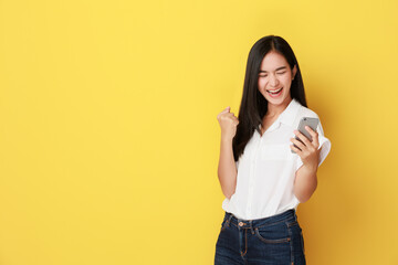 Cheerful asian woman standing isolated over yellow background, holding mobile phone, celebrating