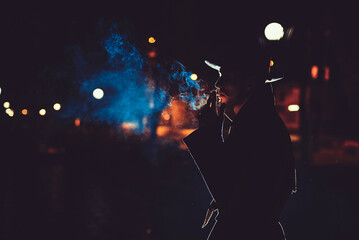 man detective agent in a hat and raincoat smokes a cigarette at night in a rainy city in the style...