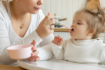 Close-up Baby girl eating blend mashed food sitting, on high chair, mother feeding child, hand with spoon for vegetable lunch, baby weaning, first solid food for kid. Healthy eating for baby humans