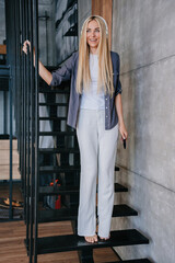 Attractive young Italian woman in headphones, grey shirt and white pants stands at stairs toothy smiles holding phone at home. Gorgeous Scandinavian blonde girl  with long hair listening music.